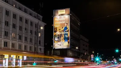 Duży podświetlony billboard na budynku nocą przedstawia chłopca w ogrodniczkach karmiącego dużego psa. Na plakacie widnieje napis: „Najlepszy przyjaciel psa? To właśnie Ty.” oraz logo marki „PsiBufet”.